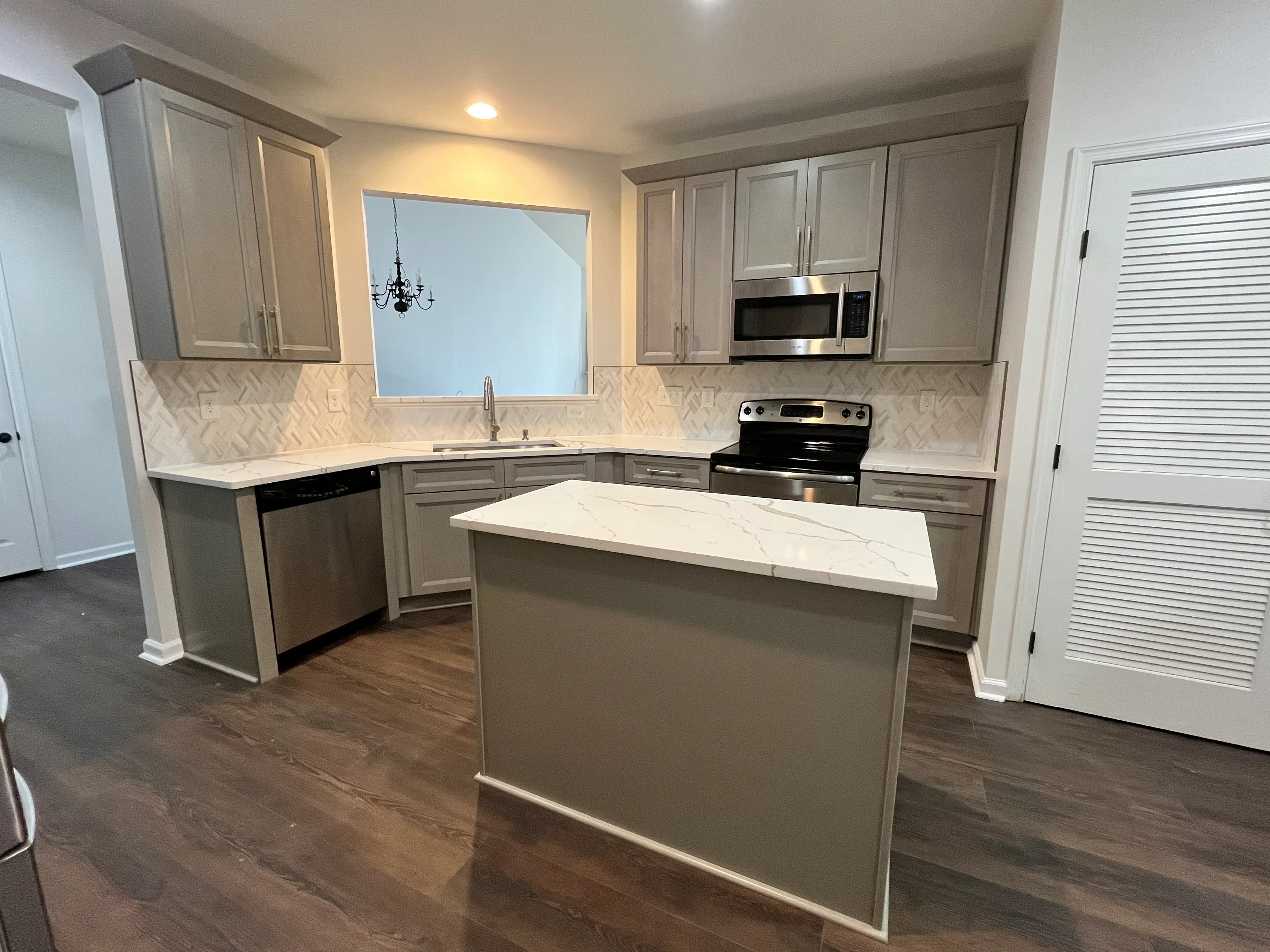 Gray cabinetry kitchen with marble countertops, island, herringbone backsplash, and stainless steel appliances